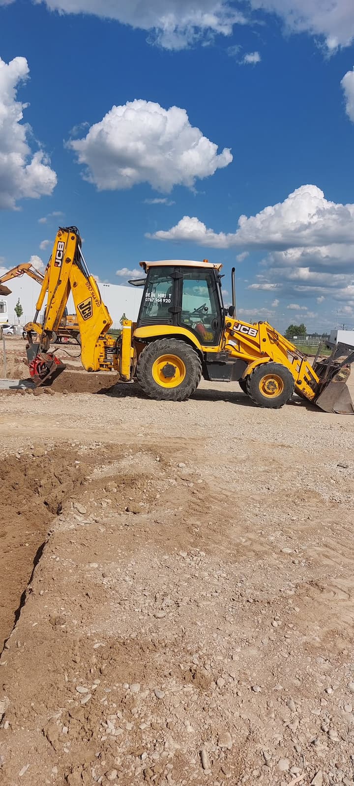 Buldoexcavator JCB cu placă compactoare pe braț, compactând umplutura unui șanț pe un șantier industrial