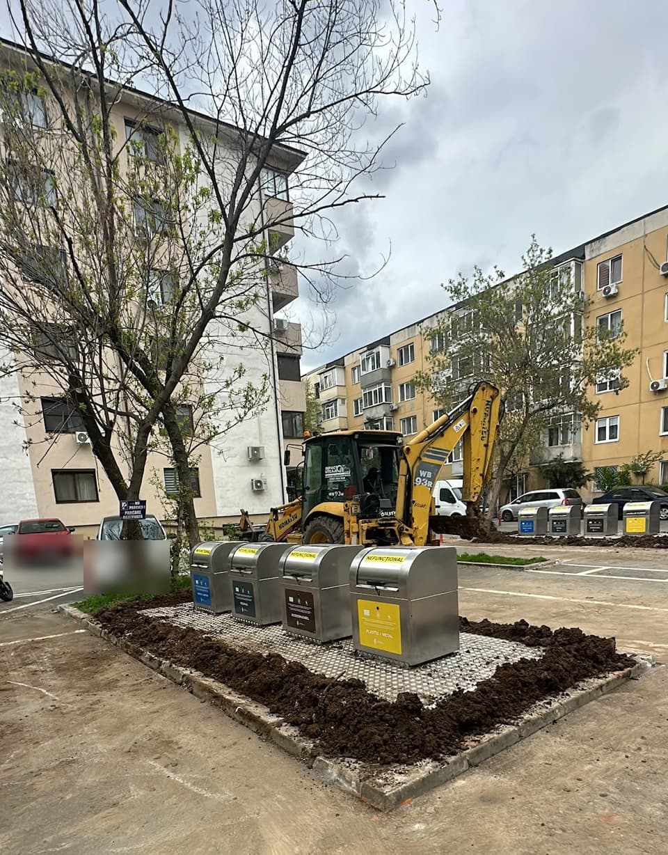 Buldoexcavator Komatsu WB93R Impact Utilaje lângă containere de gunoi semi-îngropate dintr-o parcare de bloc